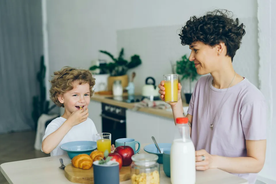 A young boy laughing at the kitchen counter with breakfast while his mother pours juice