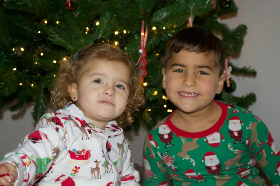 Two young children in Christmas pajamas smiling in front of a Christmas tree