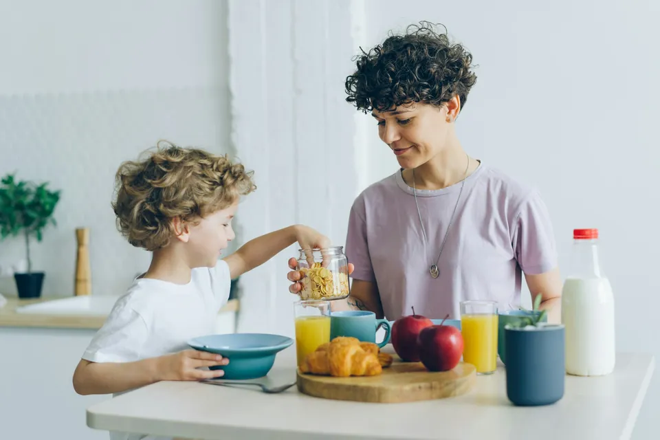 A child pouring cereal from a jar at the breakfast table while a parent watches