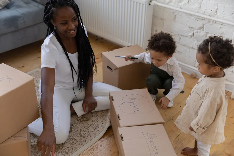 A mother and two young children packing cardboard moving boxes on the floor