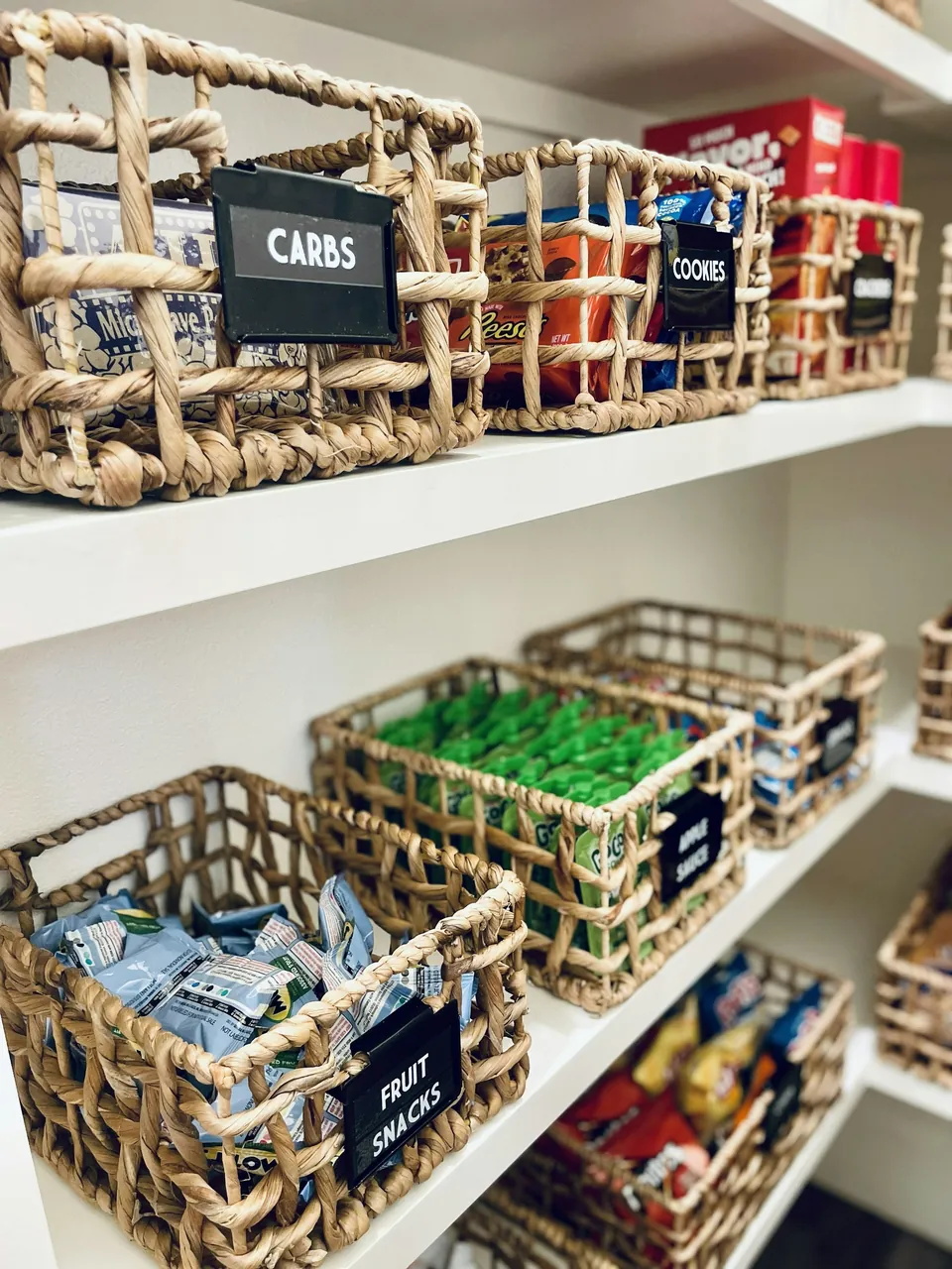 Labeled woven baskets holding snacks on white pantry shelves