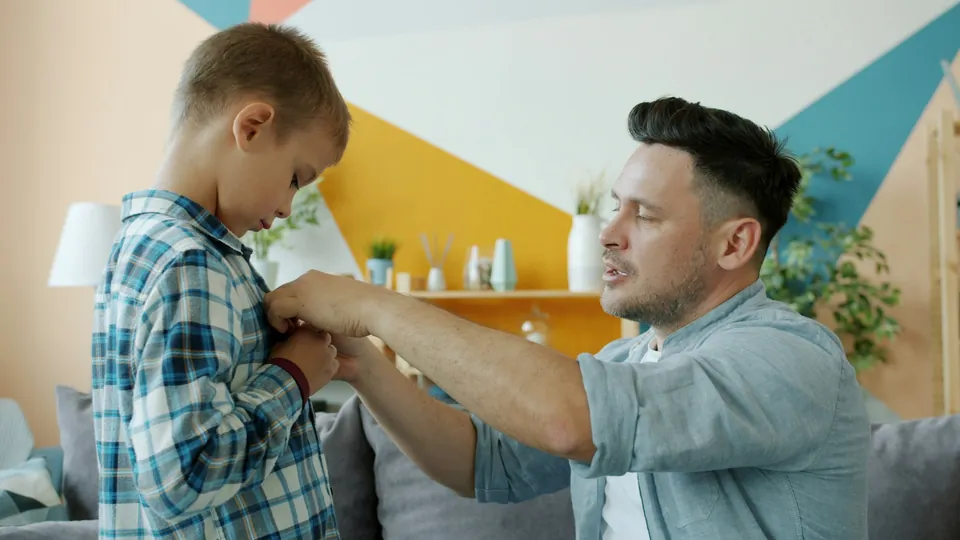 A father helping his son button his shirt in the living room