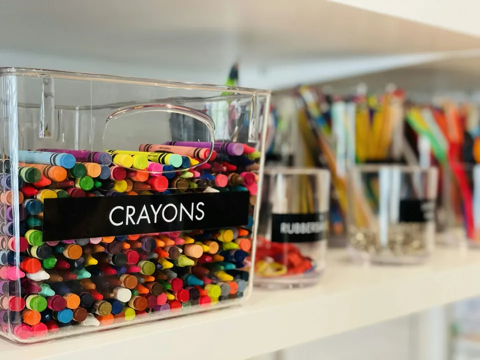 A clear bin labeled crayons on a shelf beside other art supplies