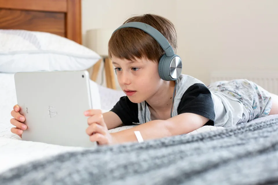 A boy wearing headphones lying on his bed using a tablet