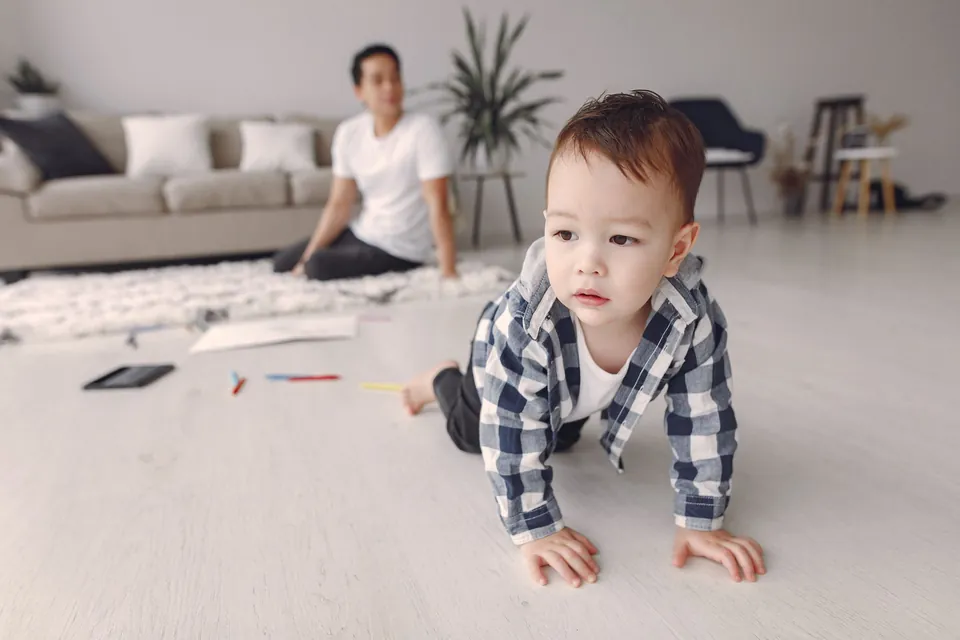 A toddler crawling across a living room floor while a parent watches from the sofa