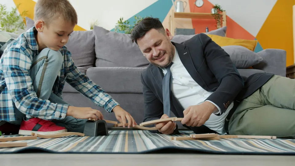 A father and son building a wooden toy train track on the living room floor